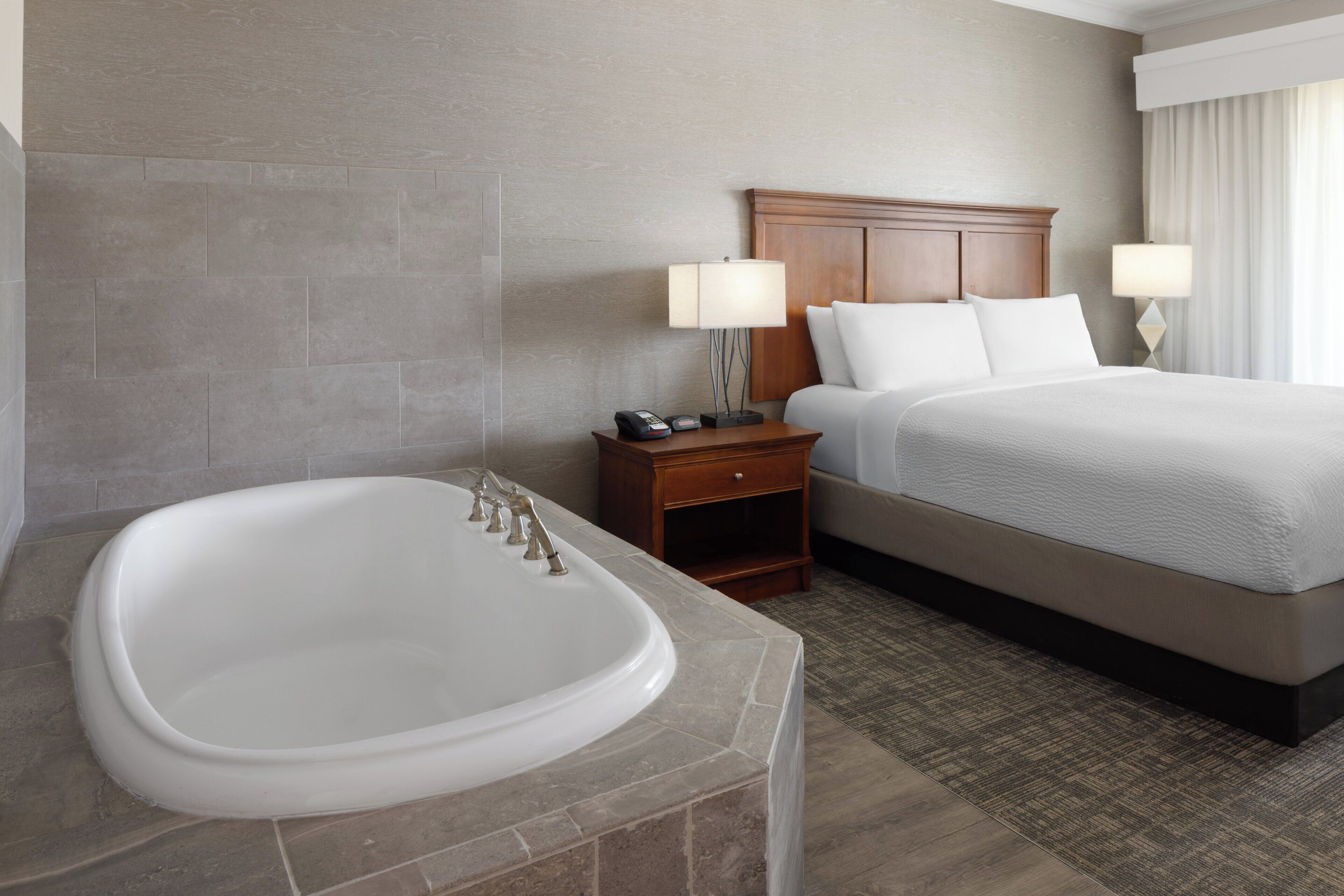 A hotel room with a large bathtub, a wooden headboard, a bed with white linens, and two bedside lamps.