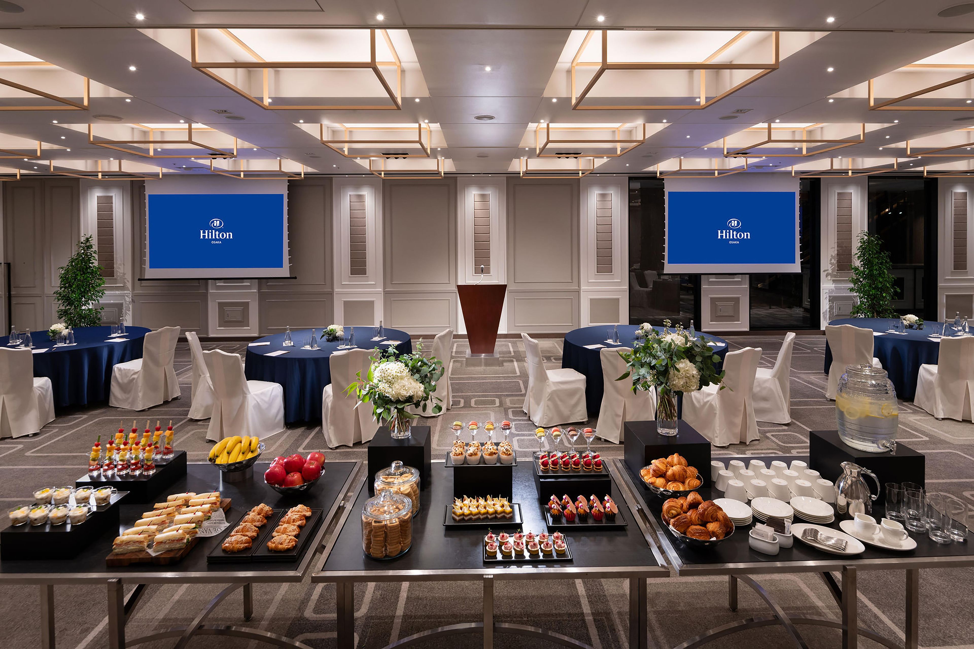 Ein Hotelkonferenzraum mit mehreren Tischen, die für eine Besprechung vorbereitet sind, inklusive eines Buffets mit verschiedenen Speisen und floralen Tischdekorationen.