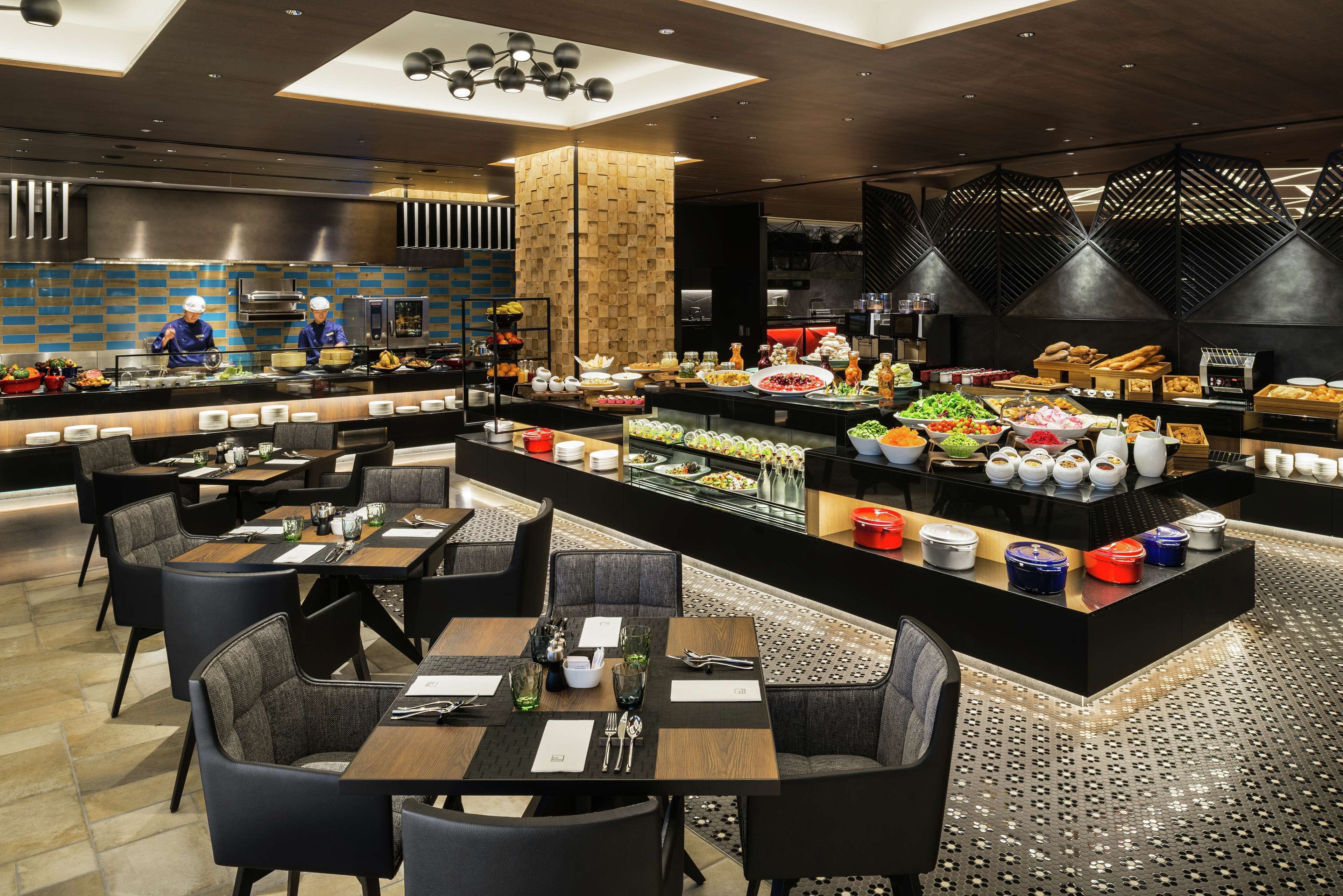 A modern restaurant with a central buffet, multiple dining tables, and a kitchen area.