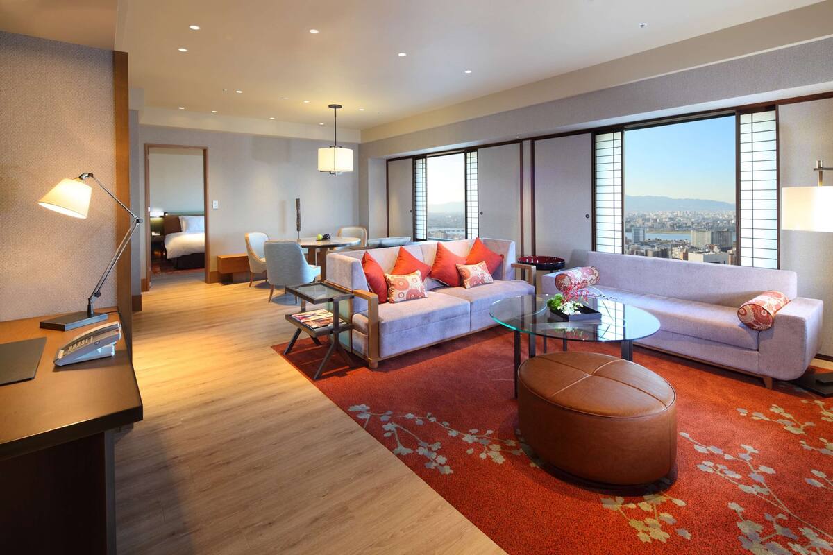 A modern hotel room with a large window, a sofa, a coffee table, and a desk with a lamp.