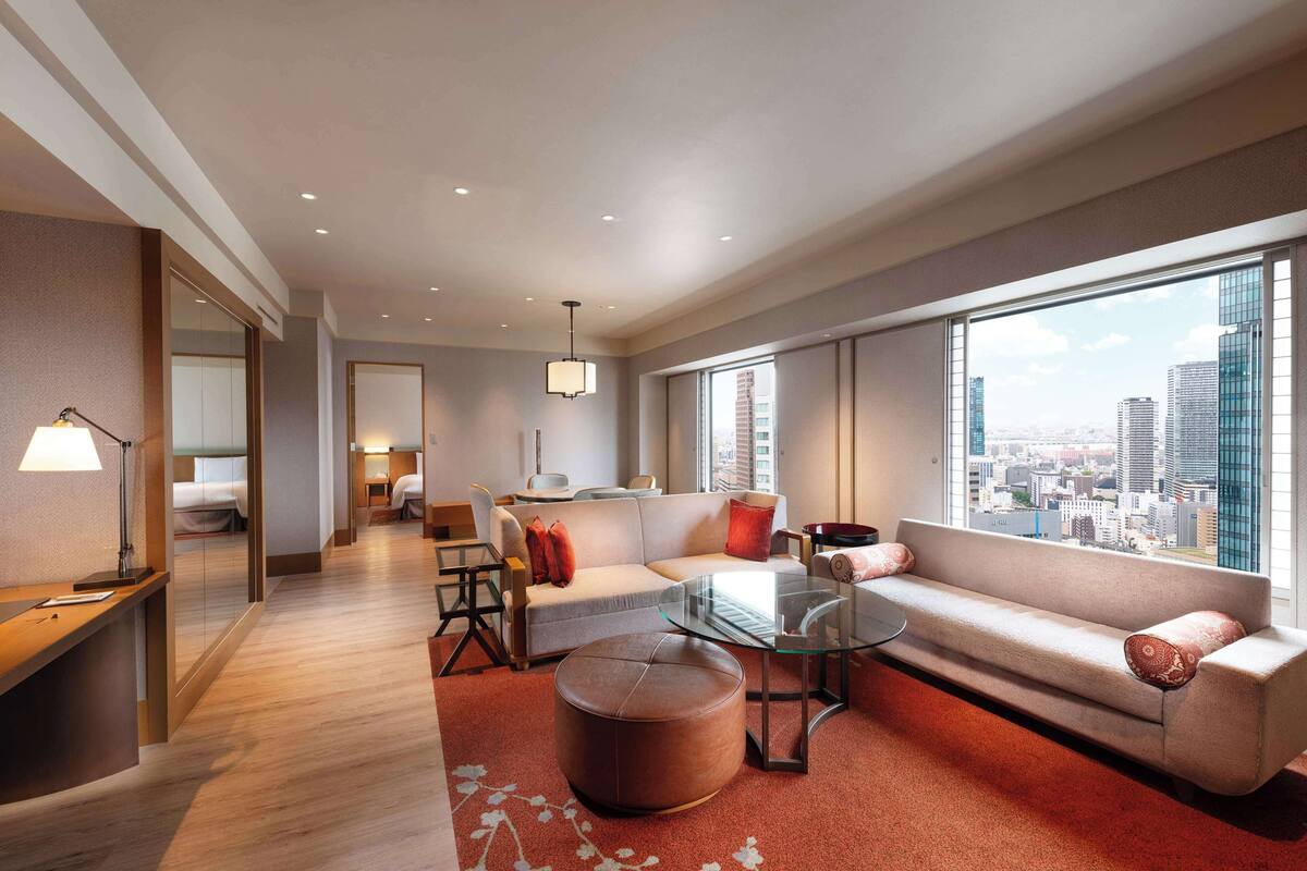 A modern hotel room with a sofa, ottoman, and glass coffee table. There is a desk with a lamp, a chair, and a bed in the background. The room has large windows with a city view.