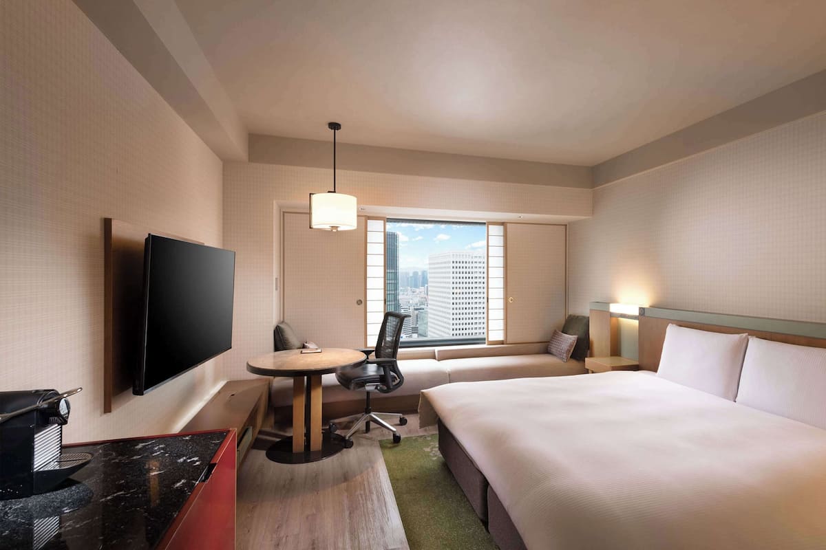 A hotel room with a large bed, a desk with a coffee machine, a TV, and a window with a city view.