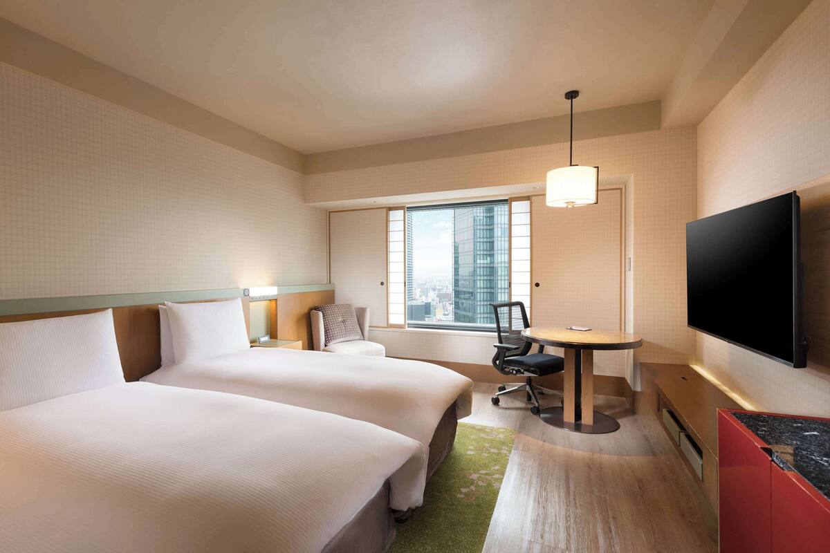 A hotel room with two beds, a desk, a chair, a TV, and a window with a city view.