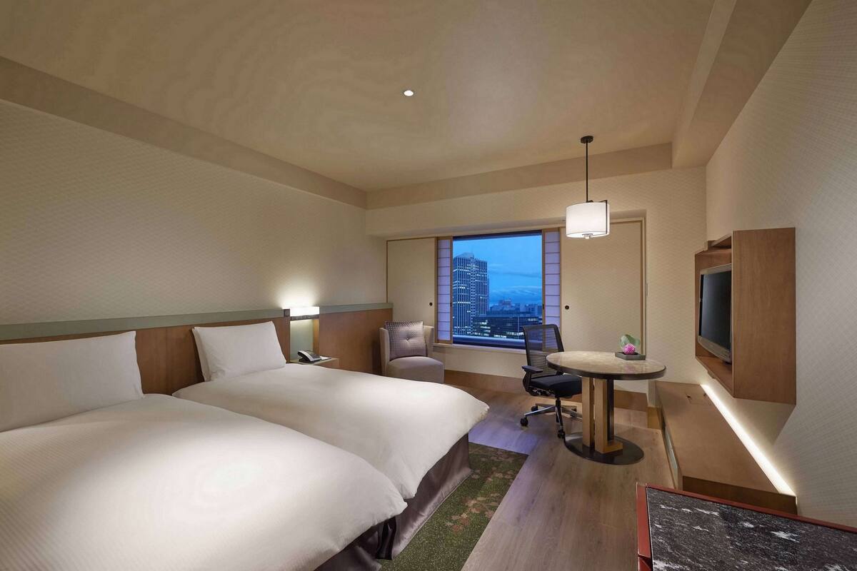 A hotel room with two beds, a desk, a chair, a TV, and a large window with a city view.
