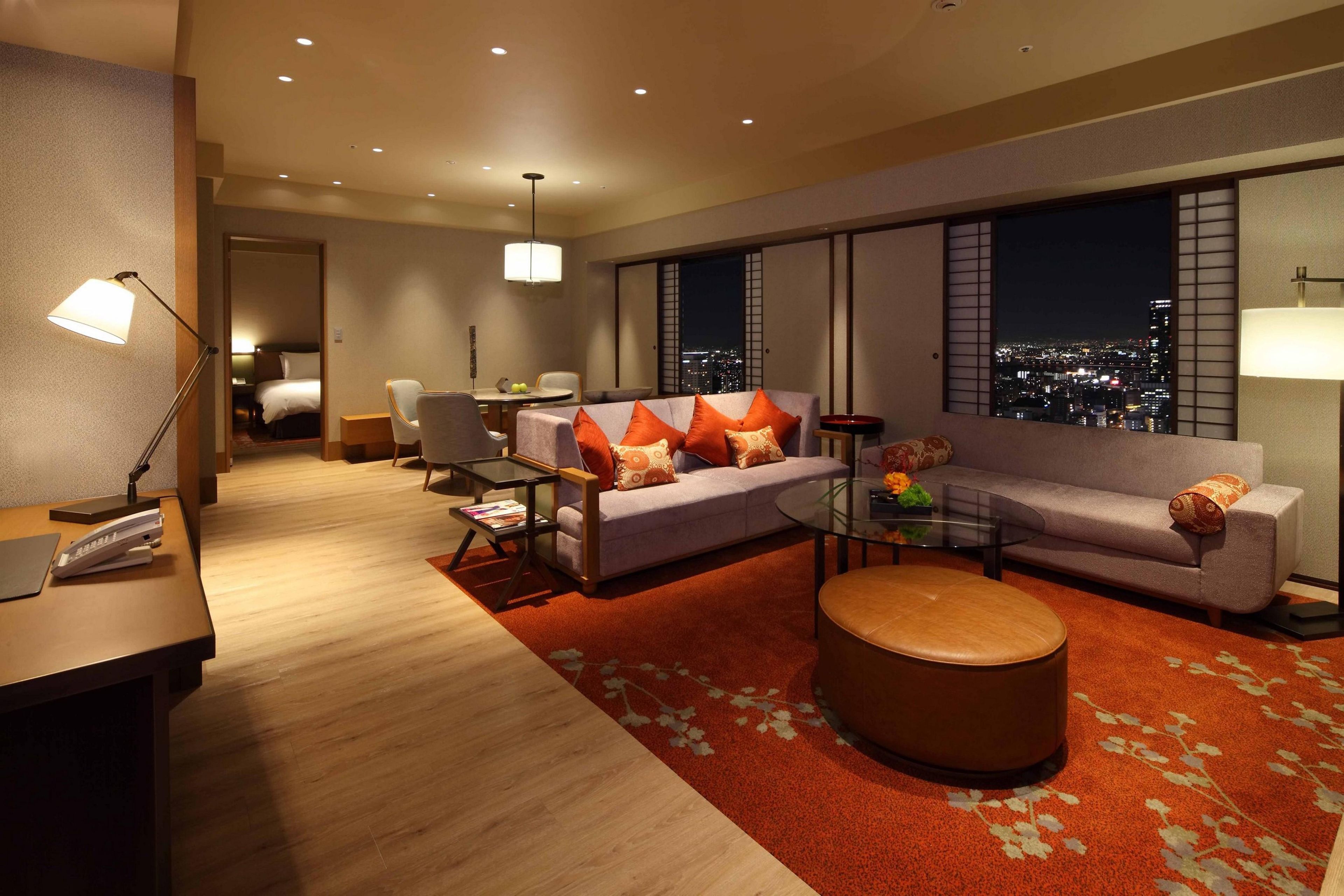 A modern hotel room with a sofa, armchairs, a coffee table, and a view of the city at night.