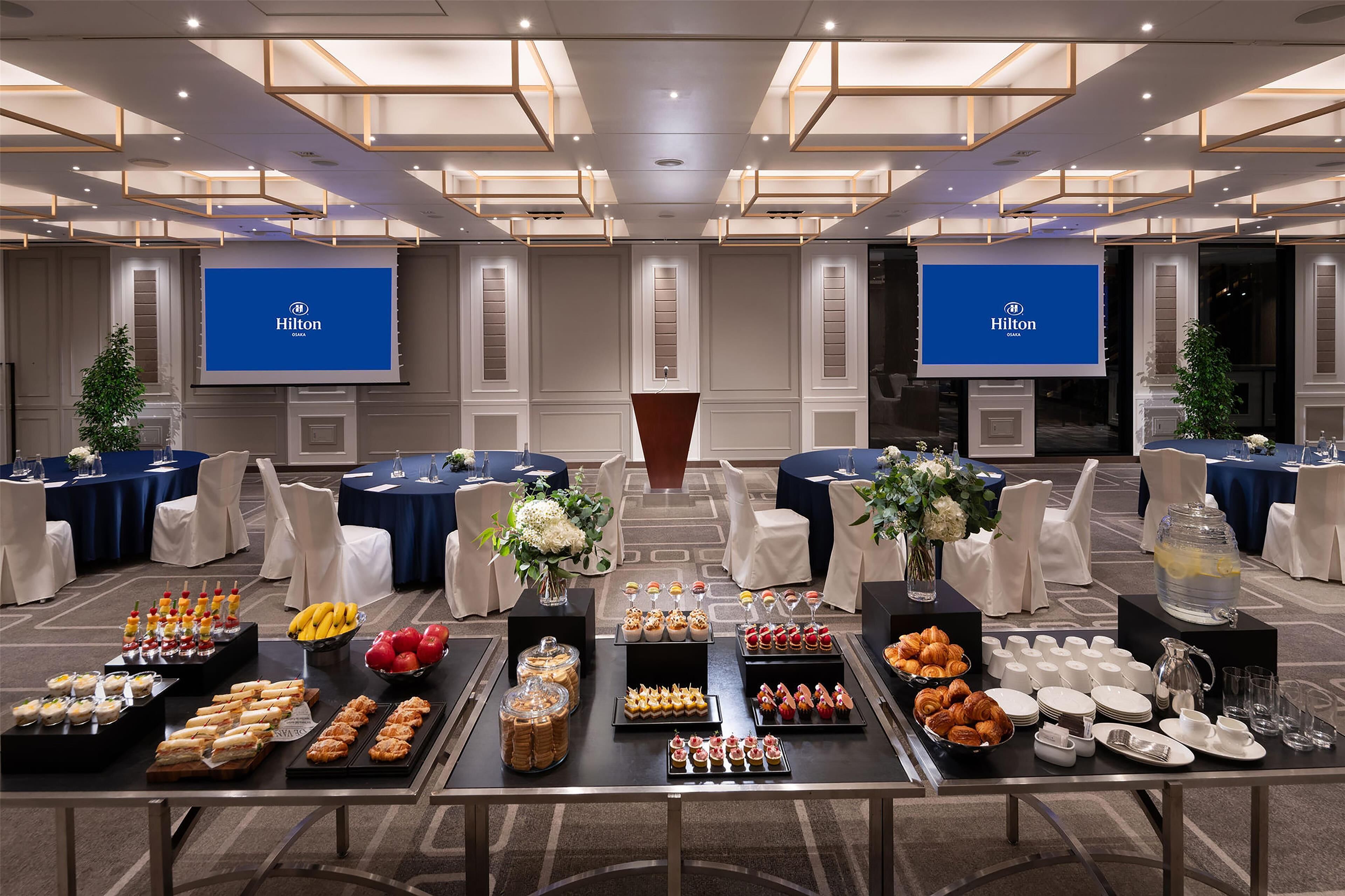 Un salón de conferencias de hotel con una mesa de buffet, múltiples pantallas mostrando el logotipo de Hilton, y una variedad de opciones de comida y bebida.
