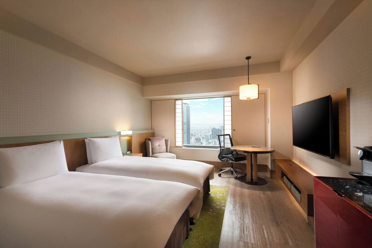 A hotel room with two beds, a desk, a chair, a TV, and a window with a city view.