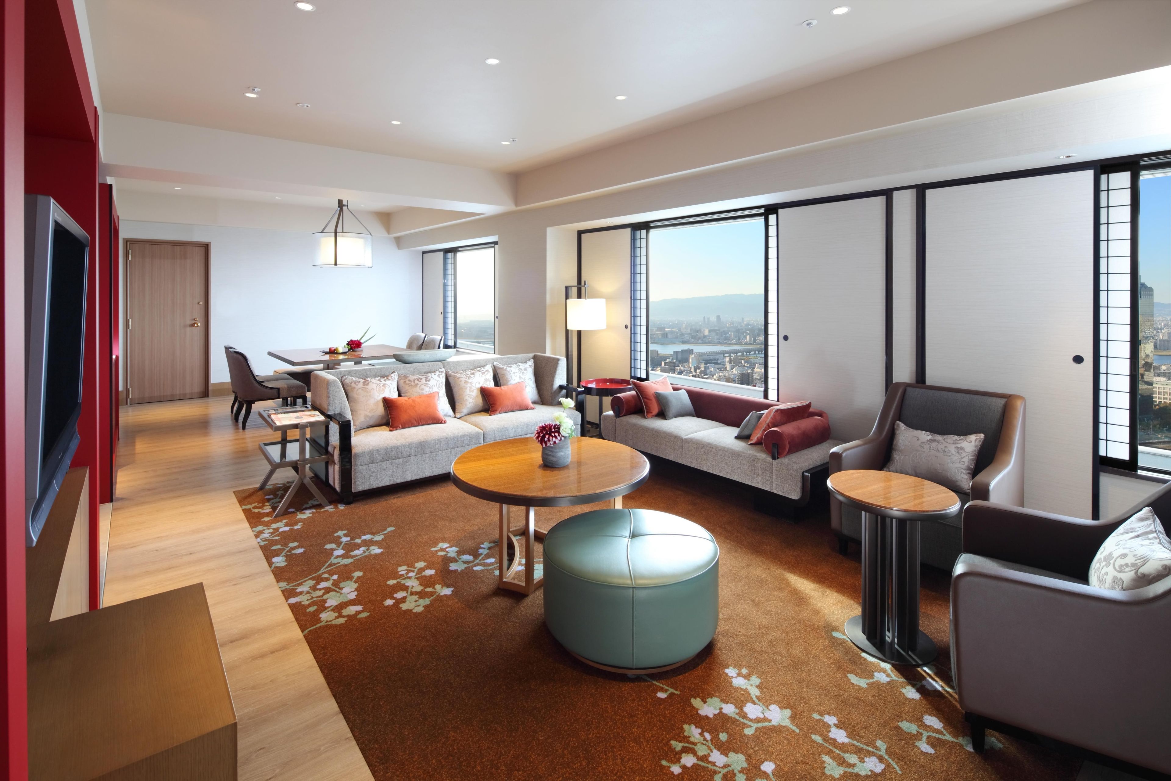 A modern hotel room with a large window, a sofa set, a dining table, and a television.
