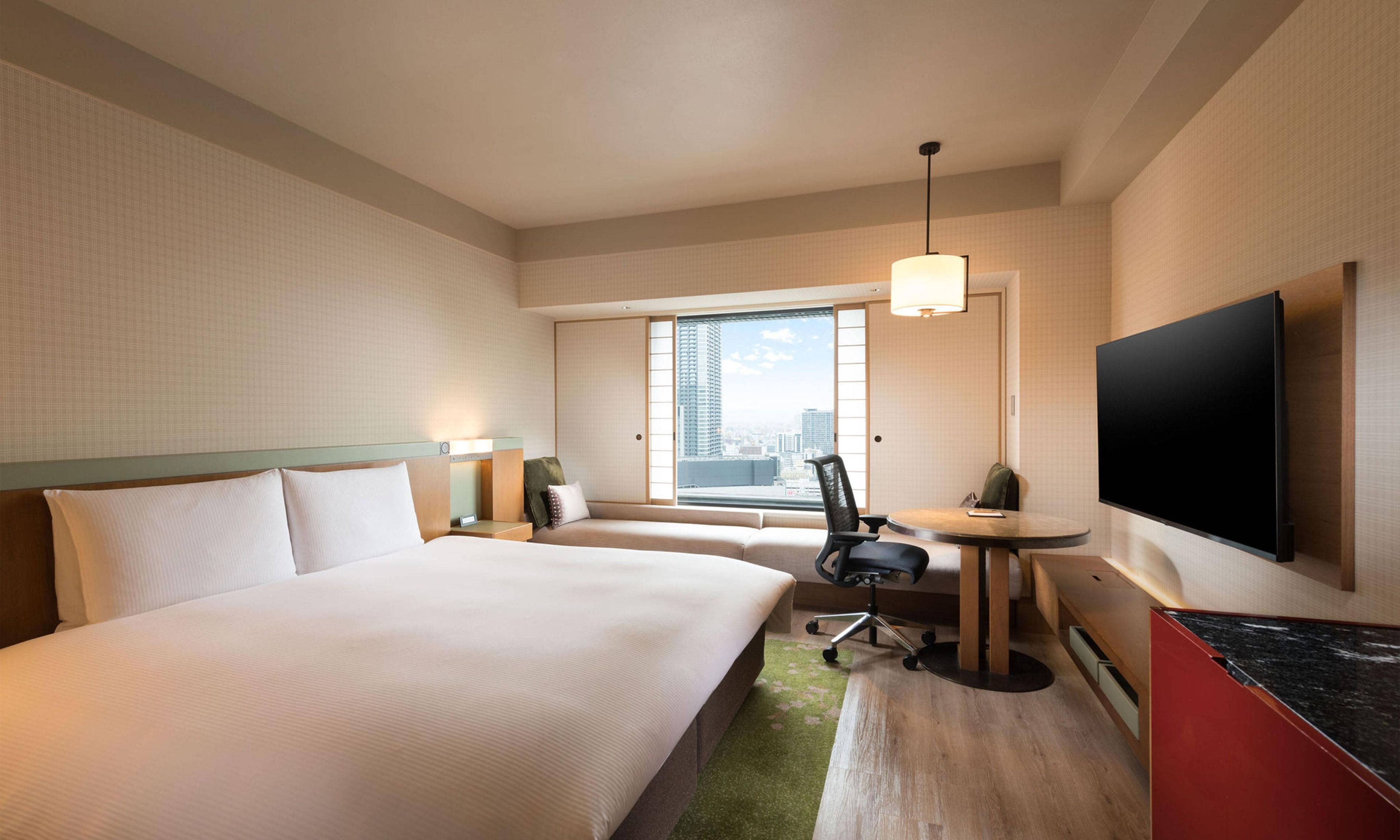 A hotel room with a bed, a desk, a chair, a TV, and a window with a city view.