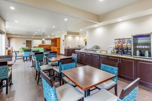 A hotel restaurant with multiple dining tables and chairs, a kitchen area, and a counter with food items.