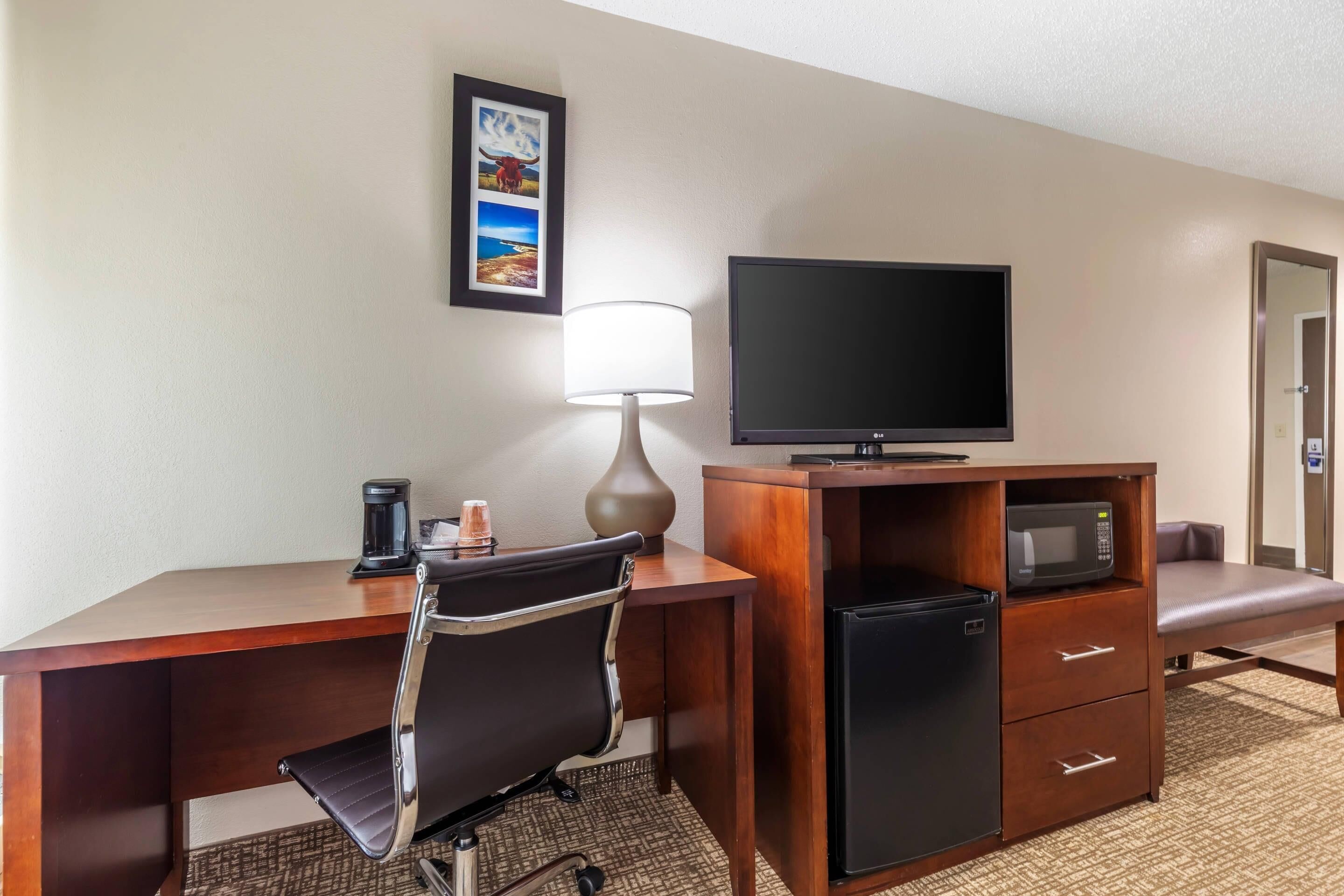 A hotel room with a wooden desk, a black office chair, a television, a refrigerator, a microwave, a lamp, and a framed picture on the wall.
