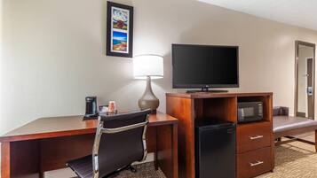 A hotel room with a wooden desk, a black office chair, a television, a refrigerator, a microwave, a lamp, and a framed picture on the wall.