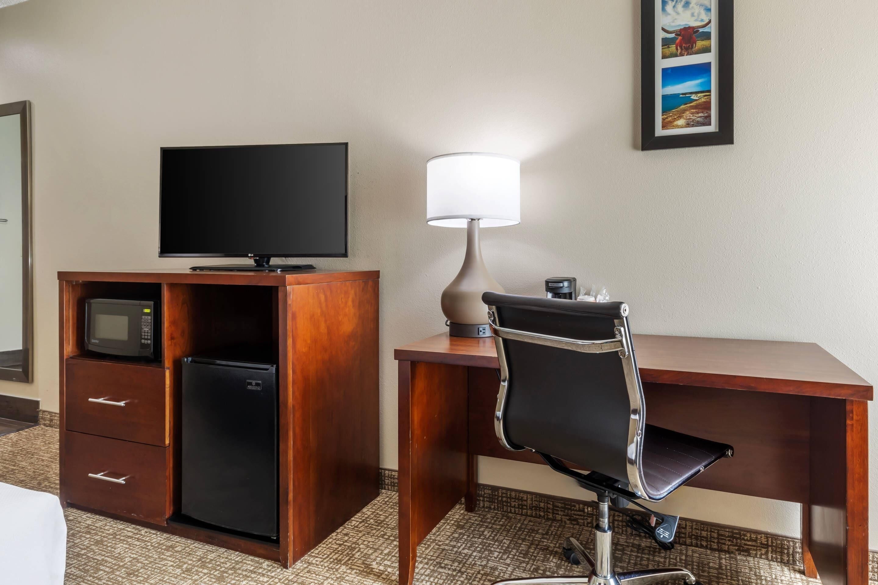 A hotel room with a wooden desk, a television, a microwave, a refrigerator, a lamp, and a chair.