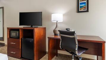 A hotel room with a wooden desk, a television, a microwave, a refrigerator, a lamp, and a chair.