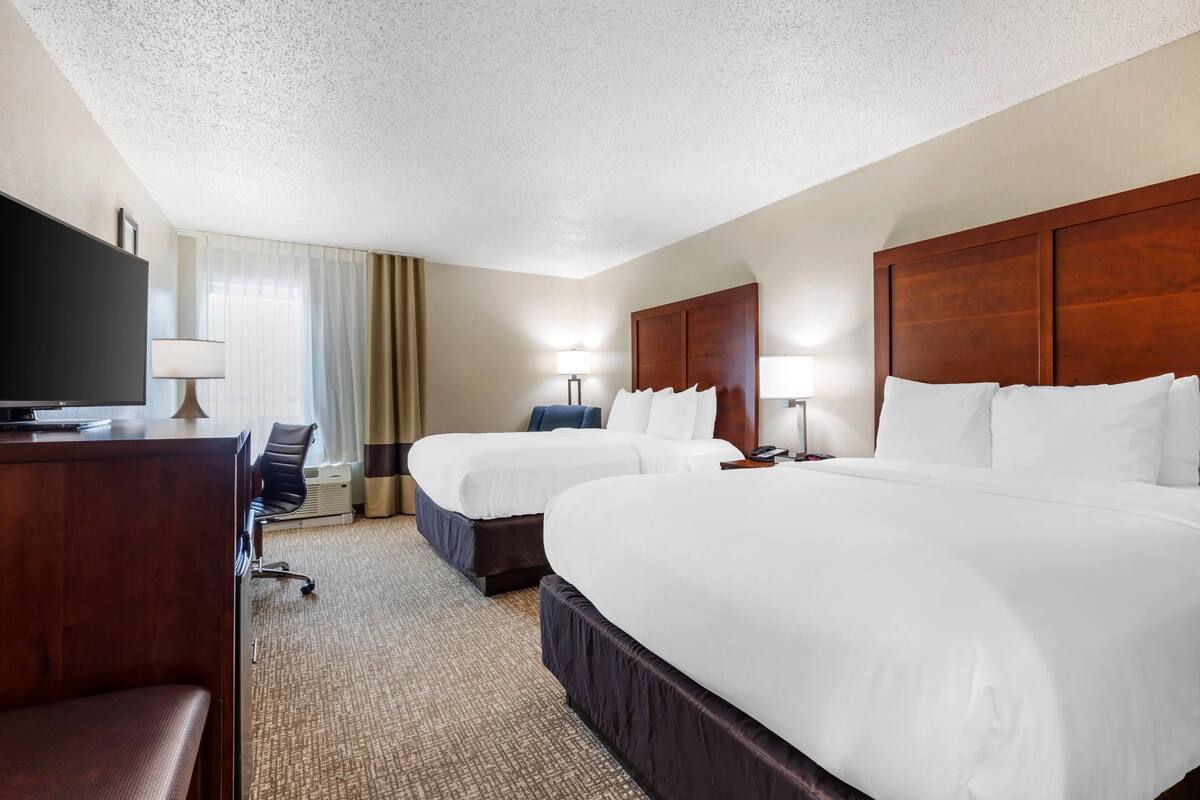 A hotel room with two beds, a desk, a chair, a TV, and lamps.