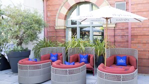 Wicker outdoor seating with red cushions and blue pillows, a white umbrella, and a brick building in the background.