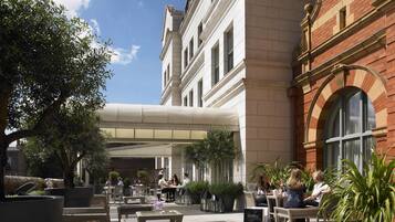 An outdoor seating area with wicker furniture and potted plants, a building with classical architecture, and a clear sky.