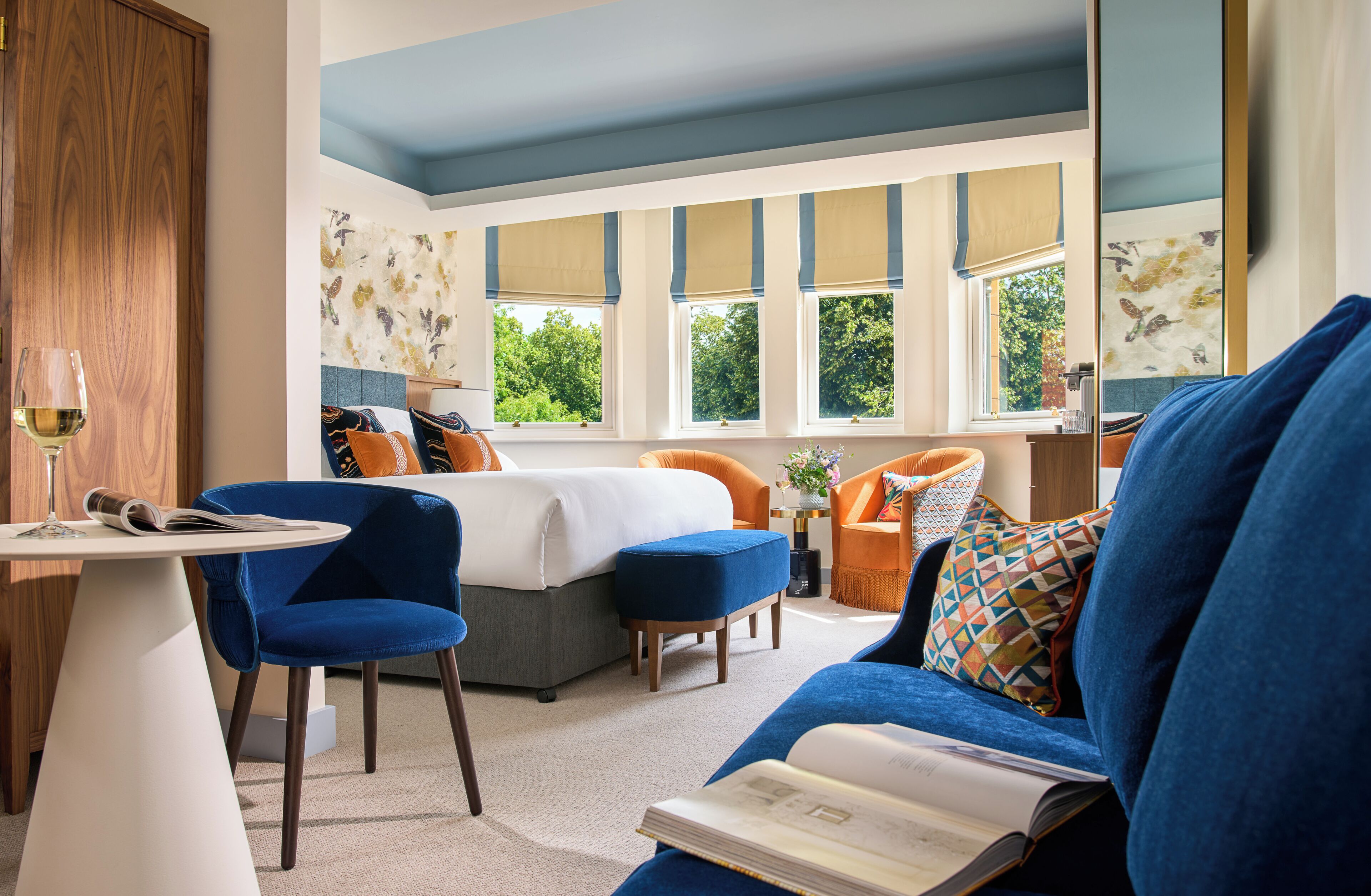 Habitación de hotel moderna con cama, sillas azules y anaranjadas, una mesita con una copa de vino y ventanas amplias con vistas a zonas verdes.