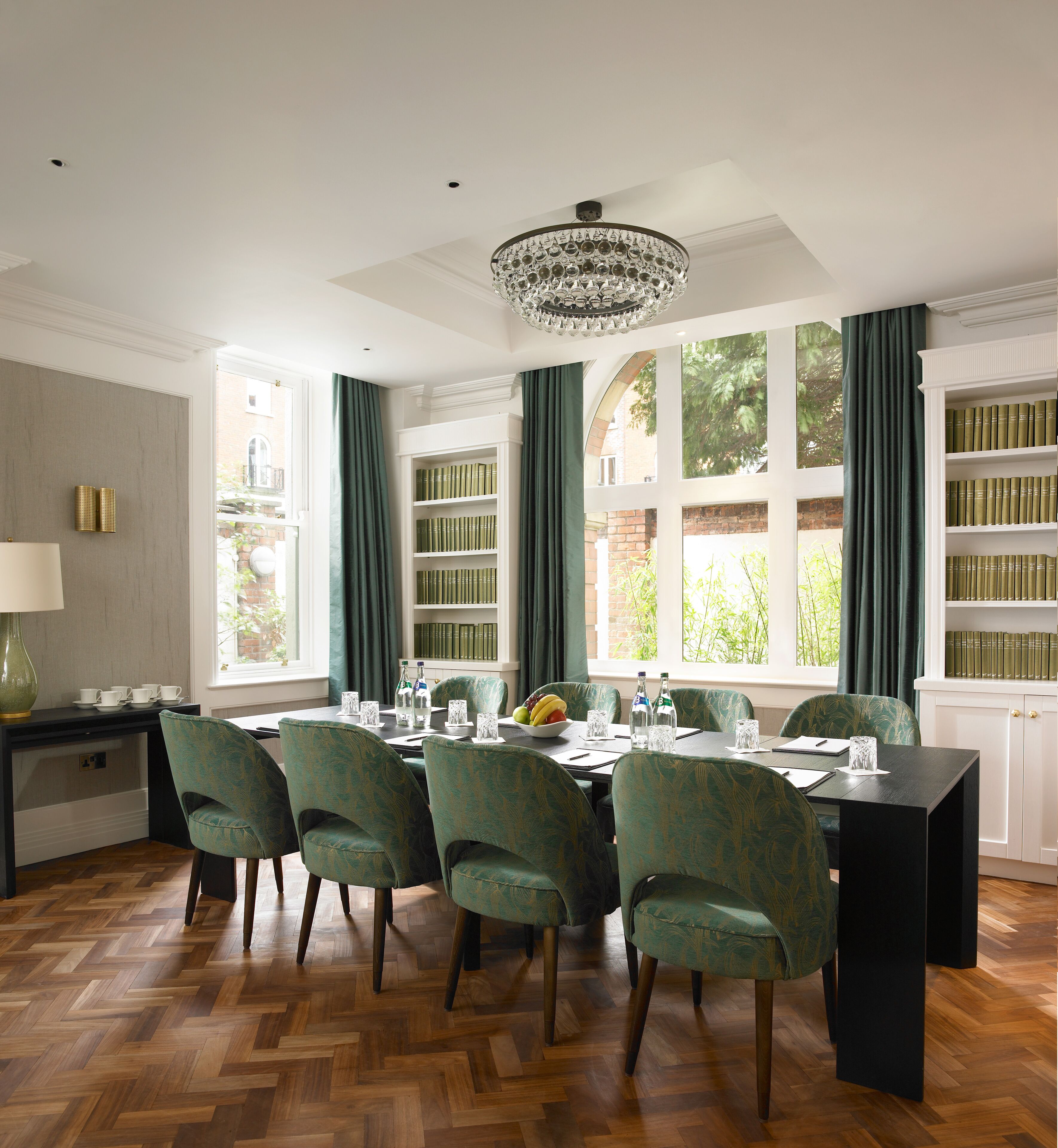 A dining area with a table set for six, surrounded by green upholstered chairs. A chandelier hangs from the ceiling, and there are bookshelves filled with books on the right.