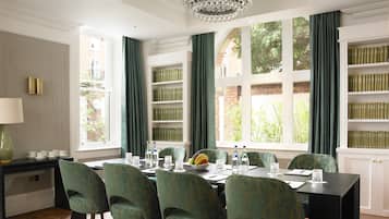 A dining area with a table set for six, surrounded by green upholstered chairs. A chandelier hangs from the ceiling, and there are bookshelves filled with books on the right.