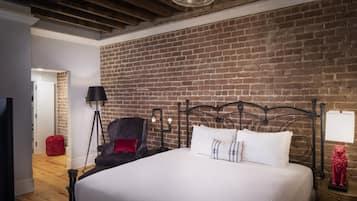 Un dormitorio con pared de ladrillo, cama con ropa blanca, una silla roja, una lámpara de pie y una estatua roja sobre una mesita de noche.