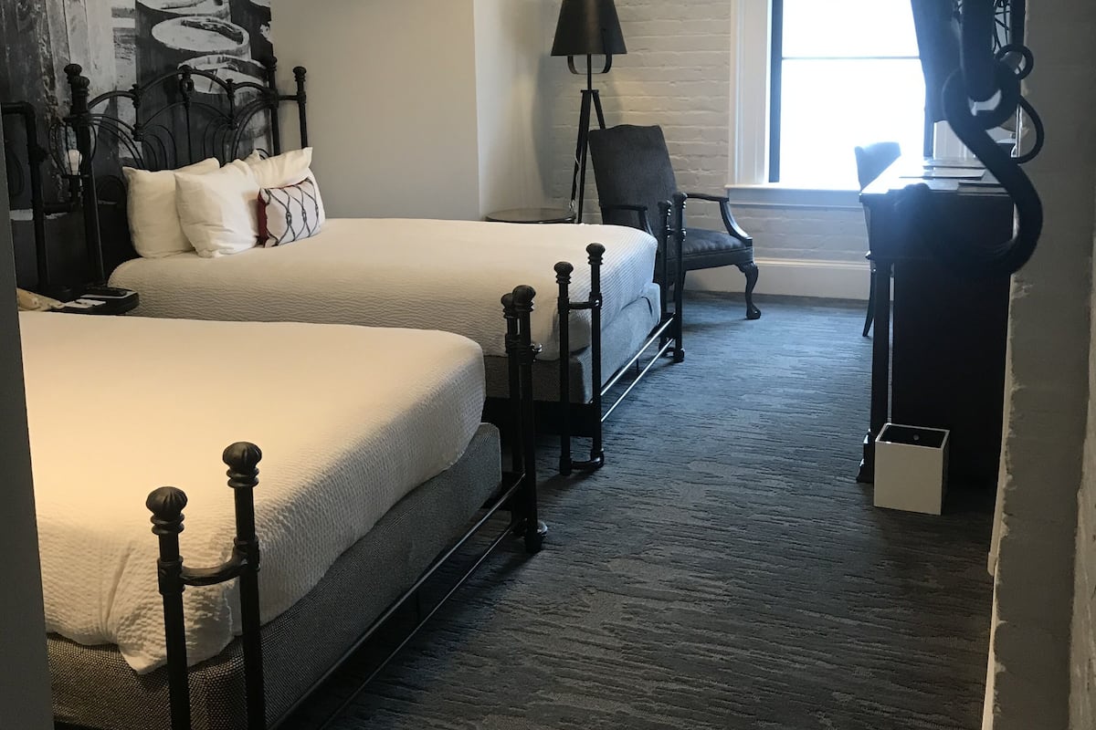 A hotel room with two beds, a desk, a chair, a lamp, and a large window.