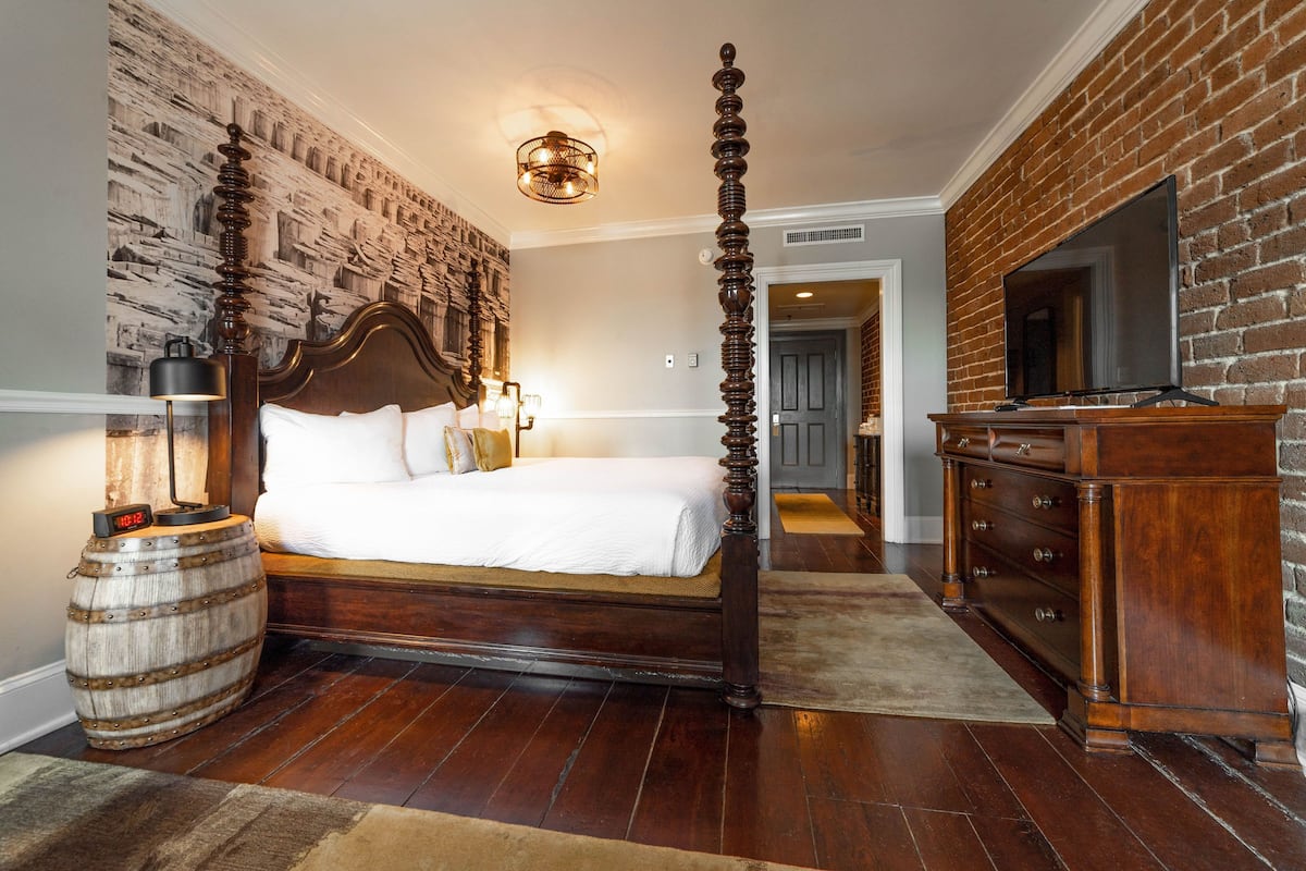 A bedroom with a four-poster bed, a wooden barrel, a brick wall, and a television on a wooden stand.
