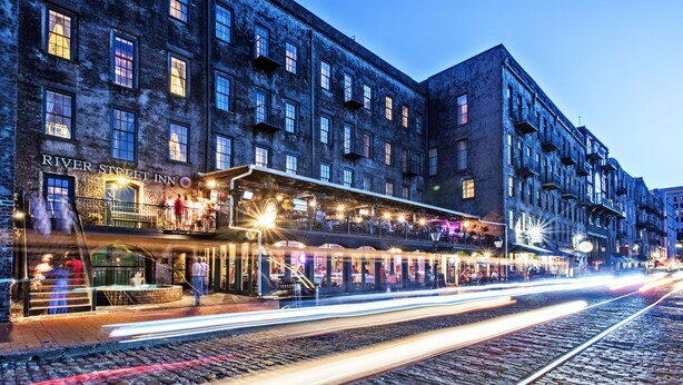 A historic building with a River Street Inn sign, featuring multiple stories, balconies, and a ground-level restaurant with outdoor seating.