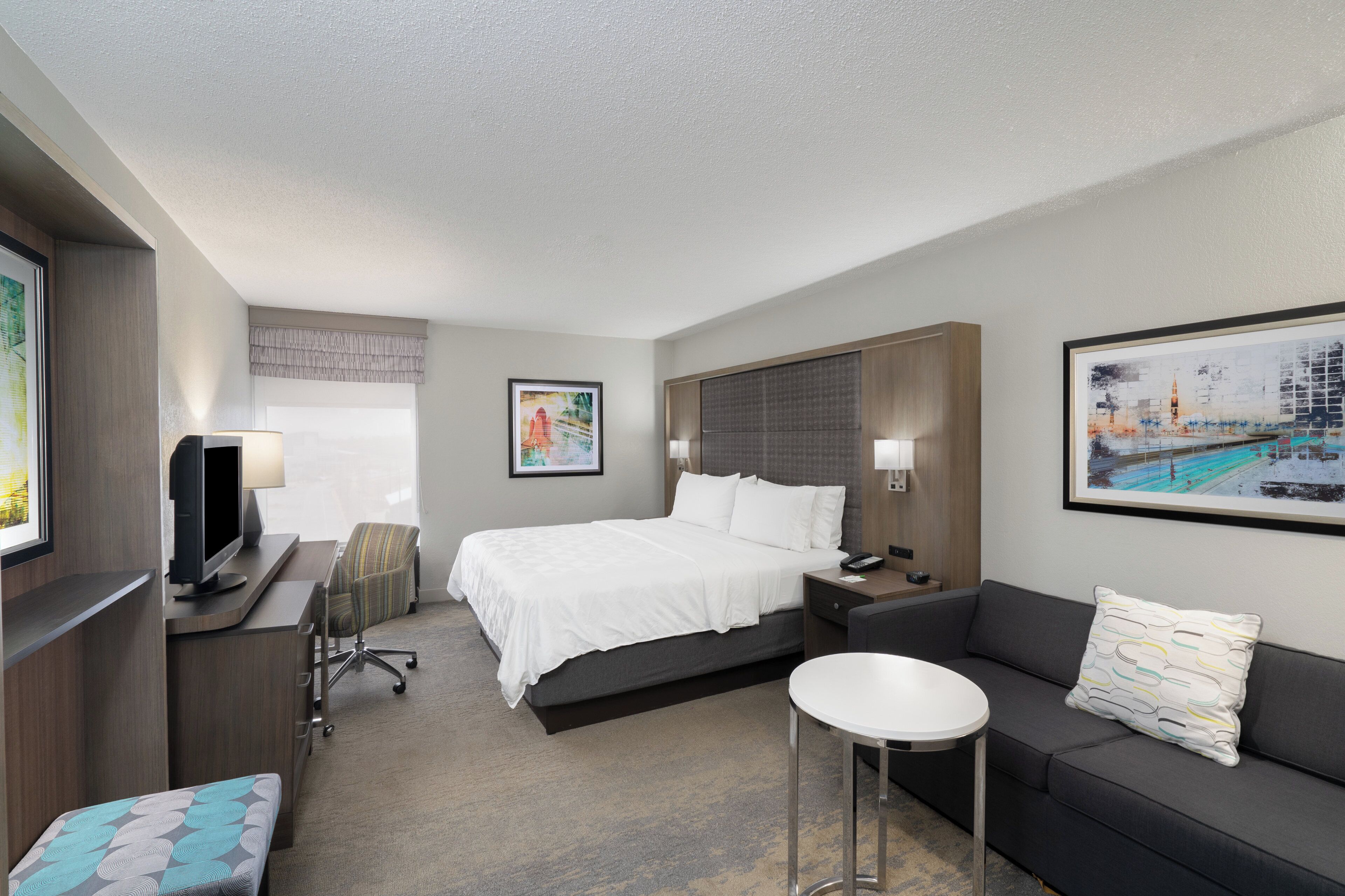 A hotel room with a bed, a desk with a computer, a TV, a sofa, a small round table, and two paintings on the walls.