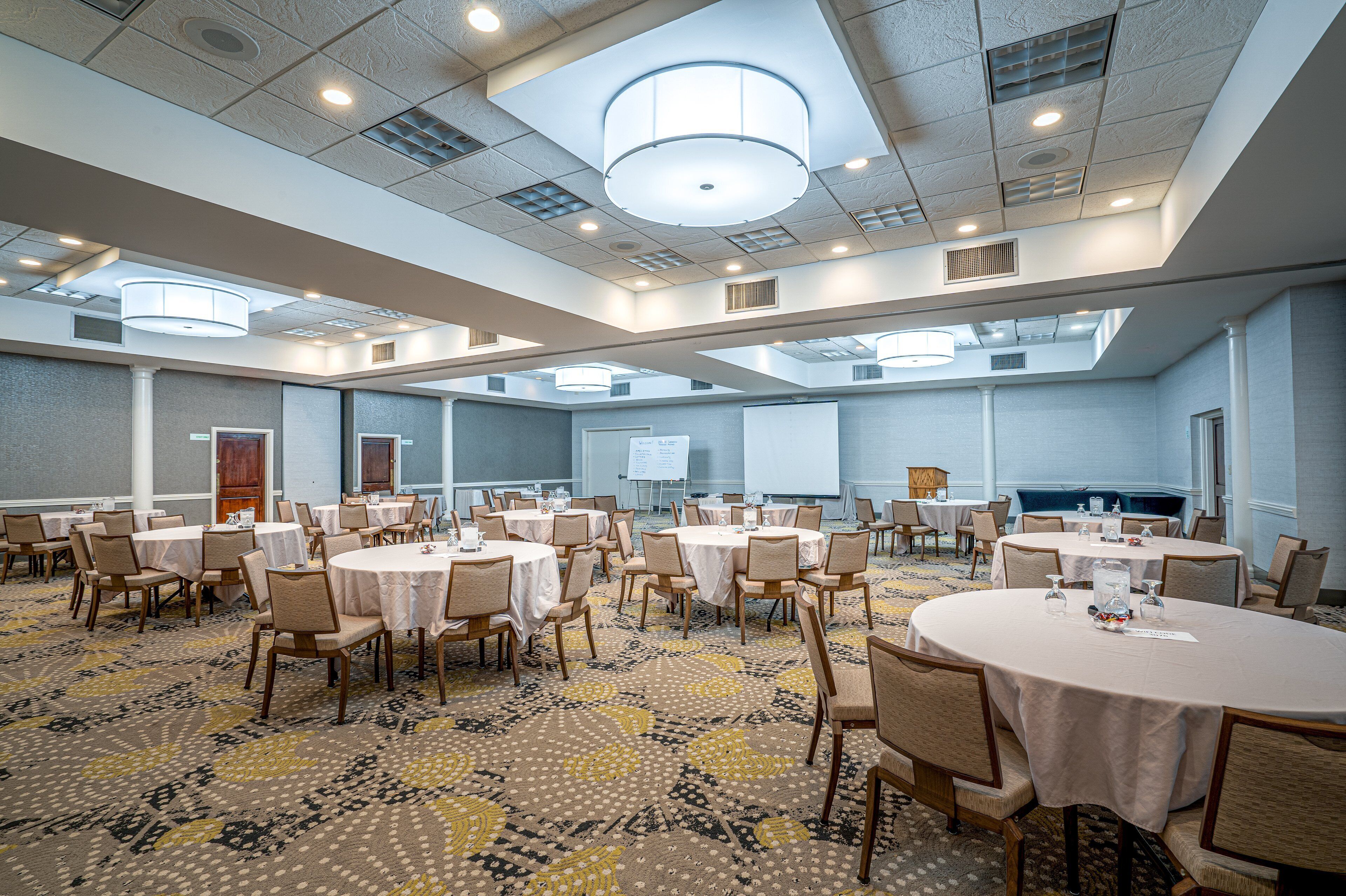 A conference room with multiple tables and chairs, a projector screen, and a presentation area.