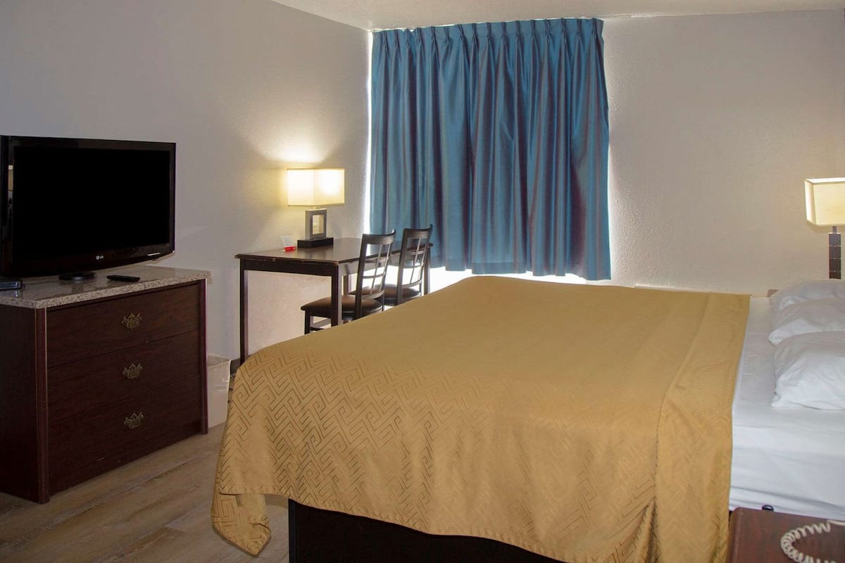 A hotel room with a bed, a television, a desk with a chair, a lamp, and a window with curtains.