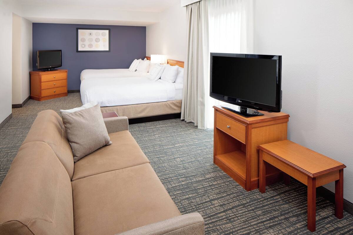 A hotel room with a bed, a TV, a wooden dresser, a sofa, and a carpeted floor.