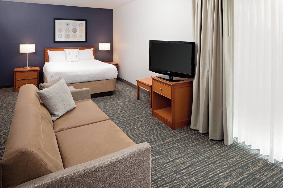 A hotel room with a bed, a TV on a wooden stand, a sofa, and a carpeted floor.