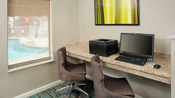 A hotel room with a desk, two chairs, a computer, a printer, and a large window overlooking a pool.