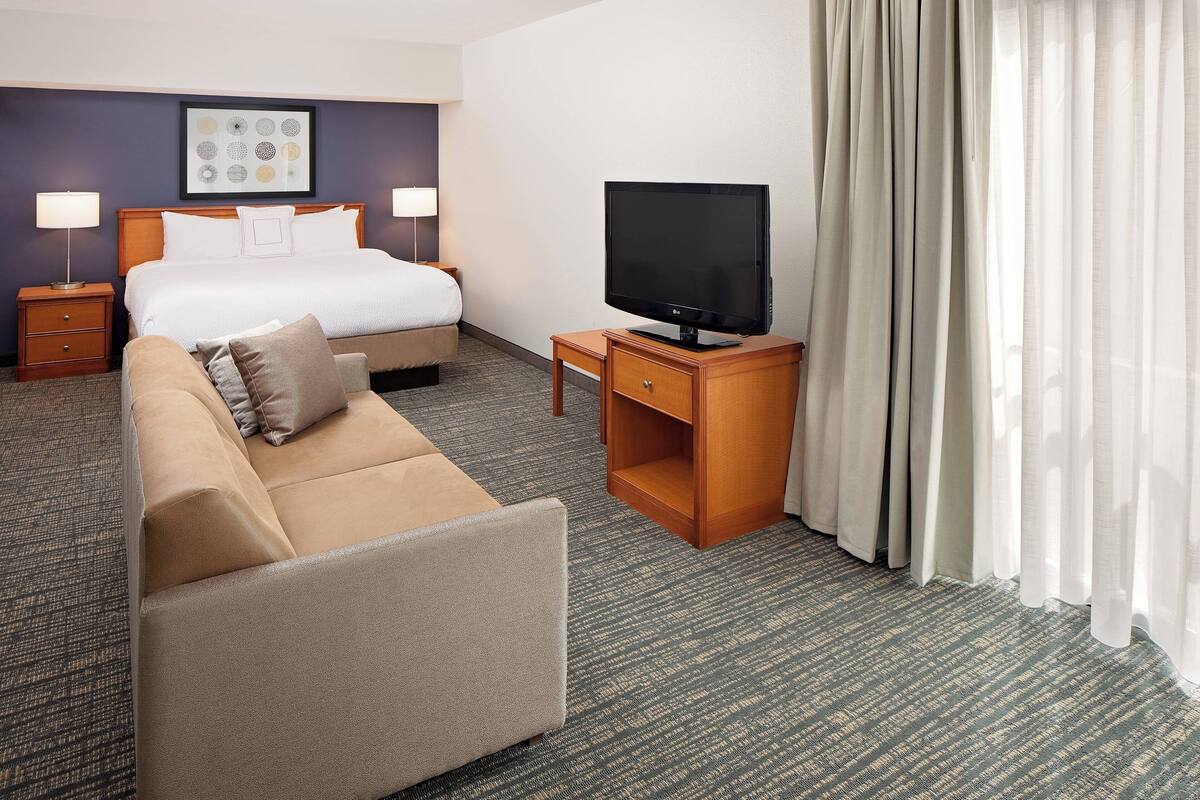 A hotel room with a bed, a TV on a wooden stand, a sofa, and a carpeted floor.