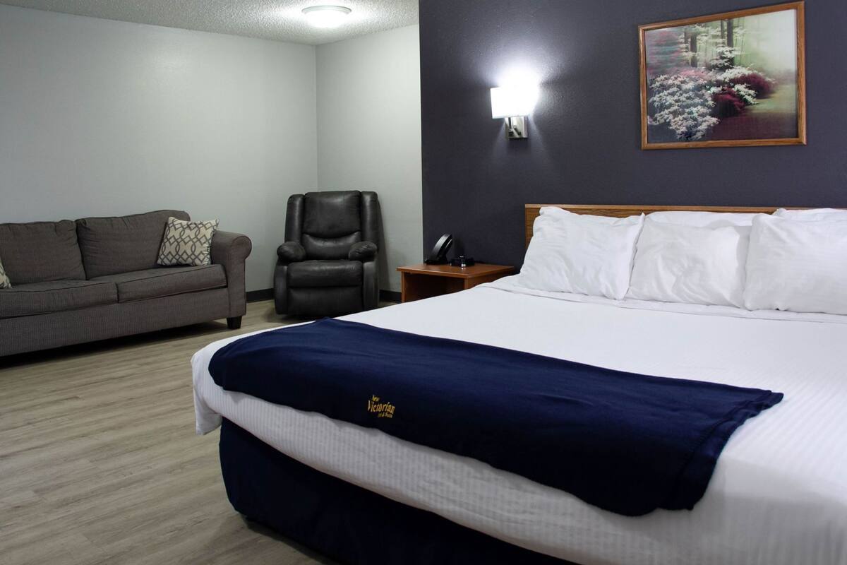 A hotel room with a bed, a sofa, a chair, a nightstand, and a wall-mounted lamp.