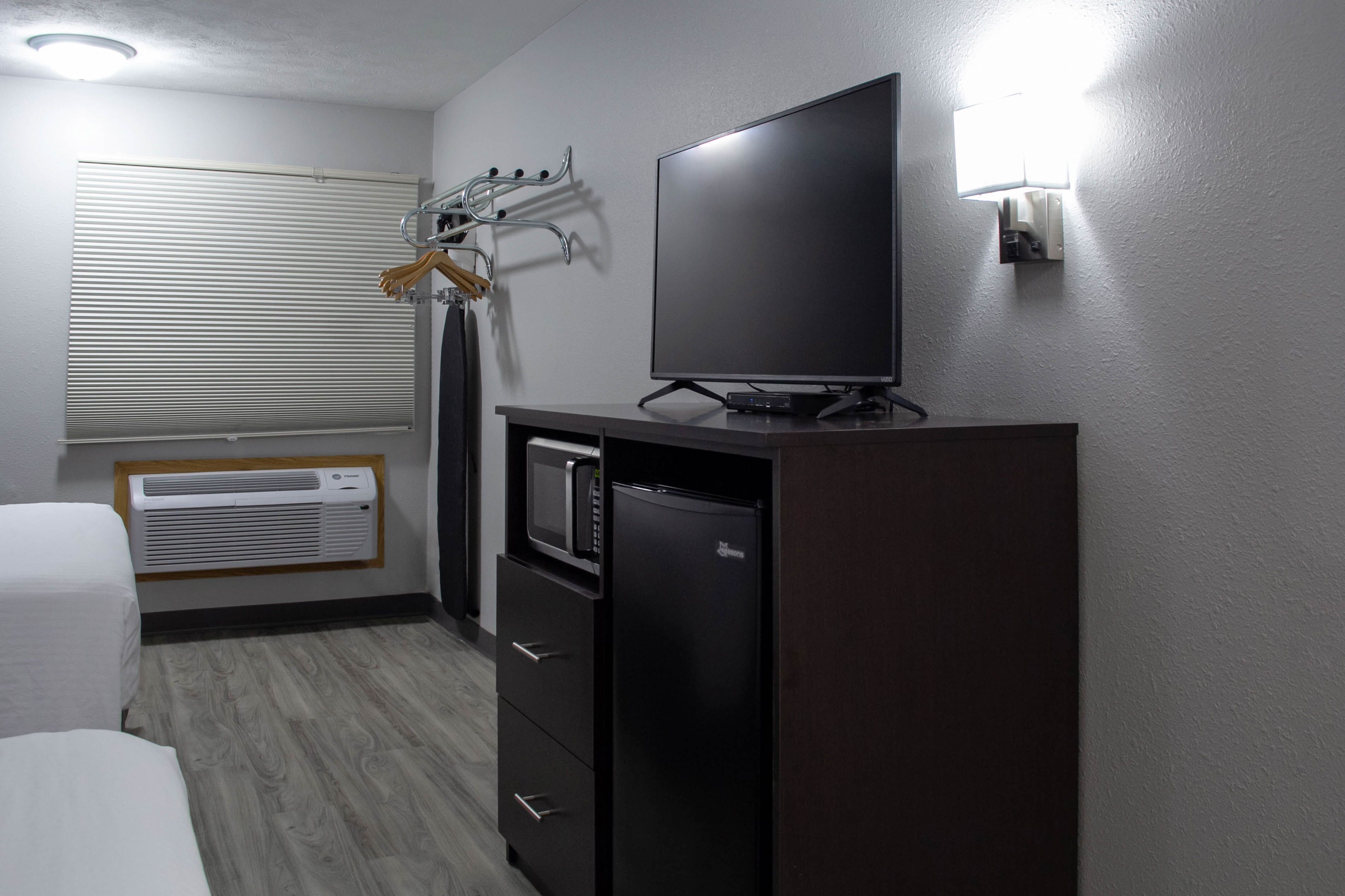 A compact hotel room with a bed, a flat-screen TV, a mini-fridge, a microwave, and a built-in heater.