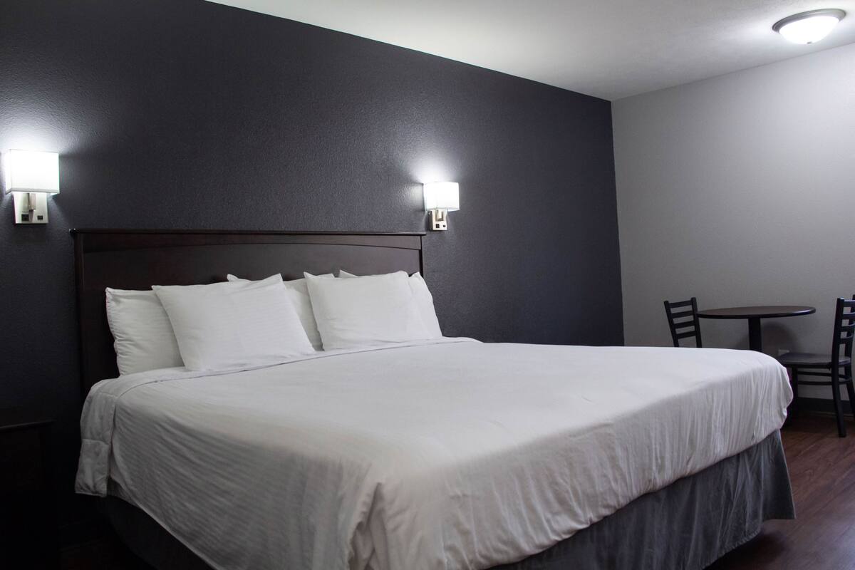 A neatly made bed with white linens in a room with dark walls and a wooden floor. There are two wall-mounted light fixtures on either side of the bed.