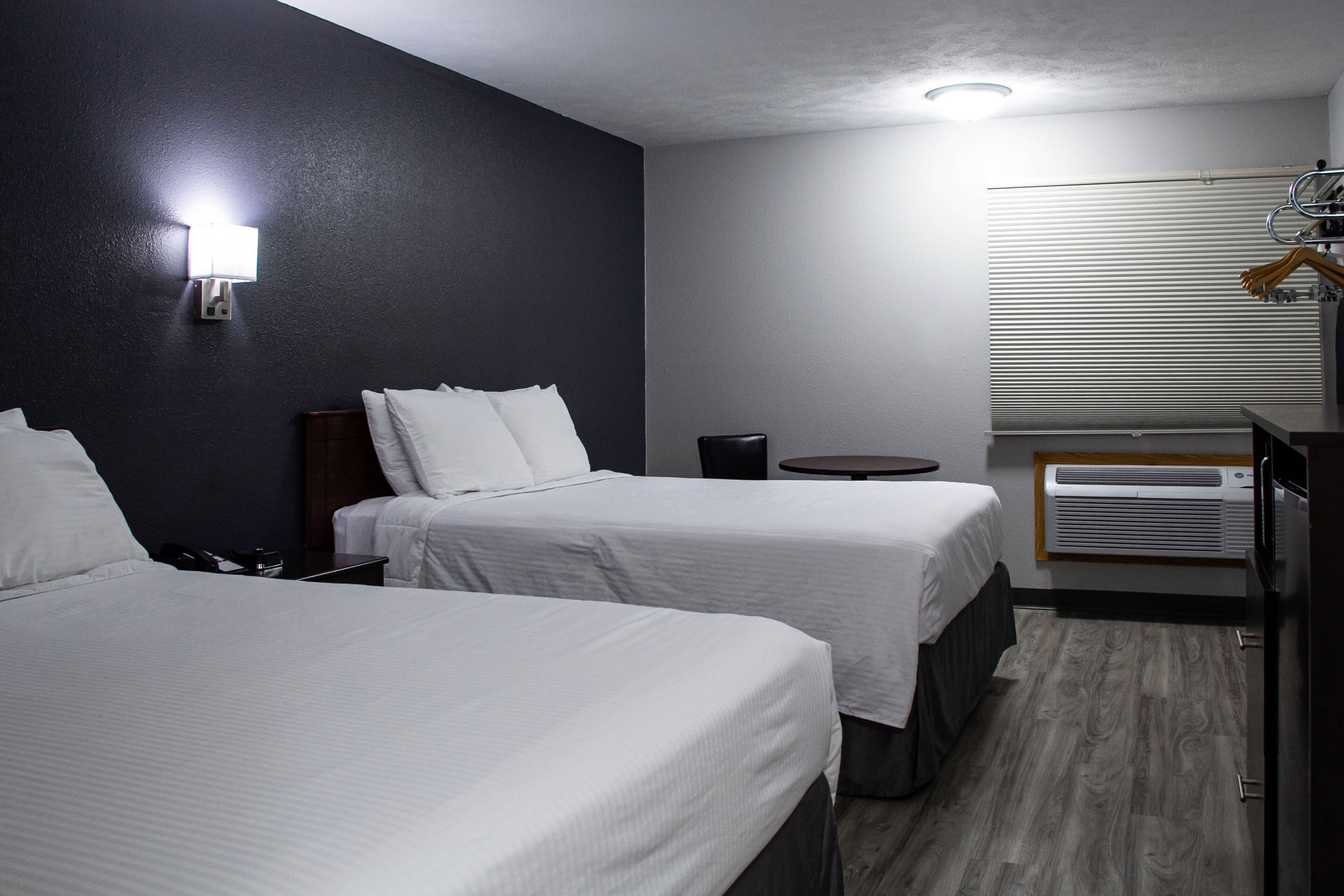 A hotel room with two beds, a small table, a chair, a desk, and a wall-mounted air conditioning unit.