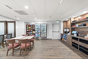 A dining area with a table and chairs, a refrigerator stocked with beverages, and shelves with packaged goods. - Hampton Inn by Hilton Los Angeles Airport LAX (Inglewood)