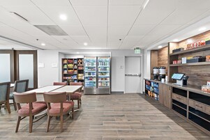 A dining area with a table and chairs, a refrigerator stocked with beverages, and shelves with packaged goods.