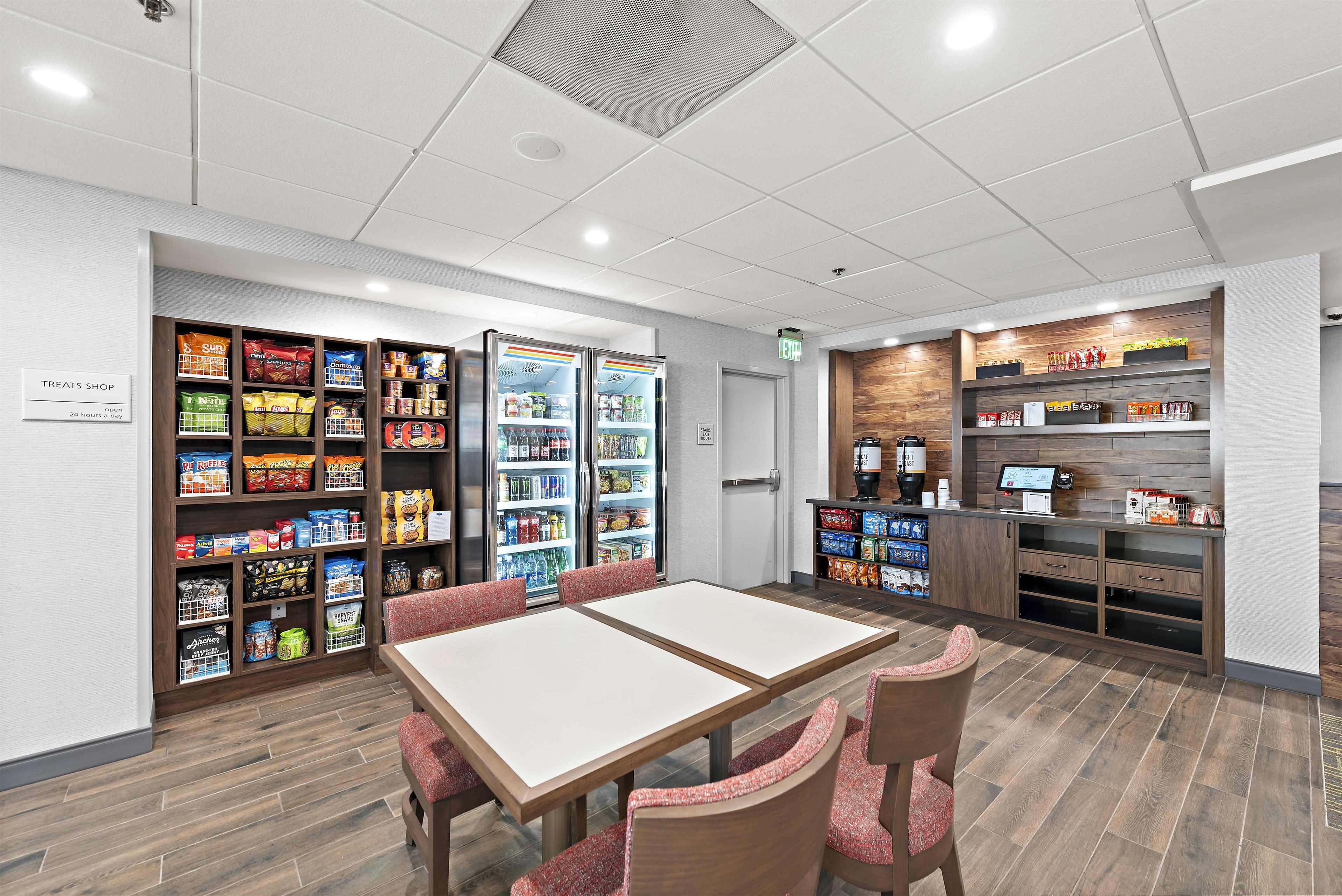 Uma loja de conveniência com diversos snacks embalados e bebidas, uma área de estar com mesas e cadeiras, e um balcão com uma máquina de café e uma tela digital.