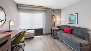 A hotel room with a wooden desk, a green office chair, a grey sofa with a red pillow, a large window with a striped blind, a wall-mounted lamp, and a framed picture on the wall. - Hampton Inn by Hilton Los Angeles Airport LAX (Inglewood)