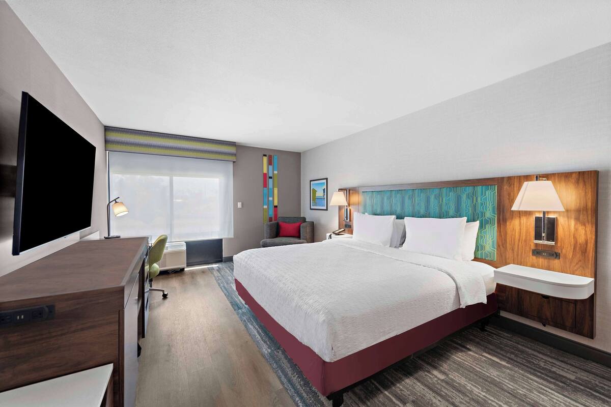 A hotel room with a large bed, a desk with a flat-screen TV, a bedside table with a lamp, and a window with a view.