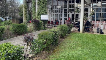 Ein Gebäude mit überdachter Außenbestuhlung, einem gepflasterten Weg und Grünflächen.