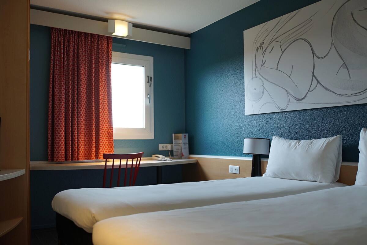 A hotel room with a bed, a desk, a chair, a window with curtains, a wall art piece, and a lamp.