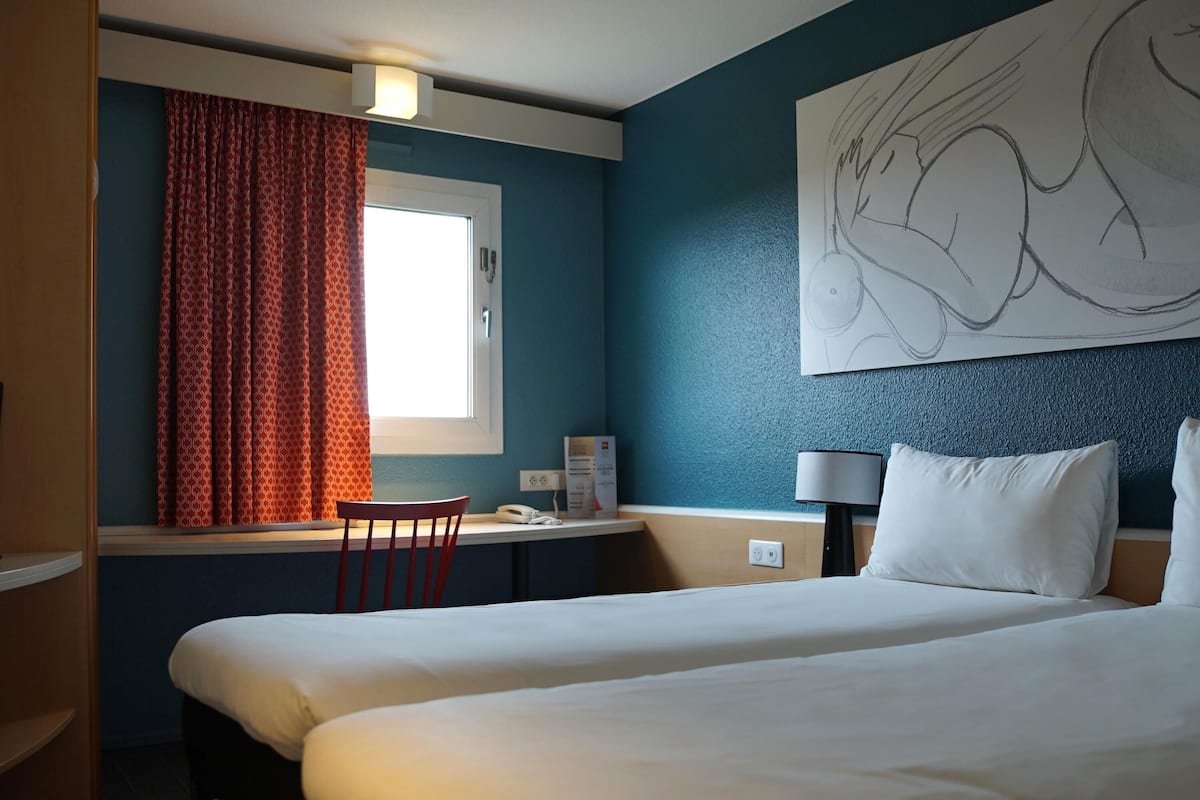 A hotel room with a bed, a desk, a chair, a window with curtains, a wall art piece, and a lamp.