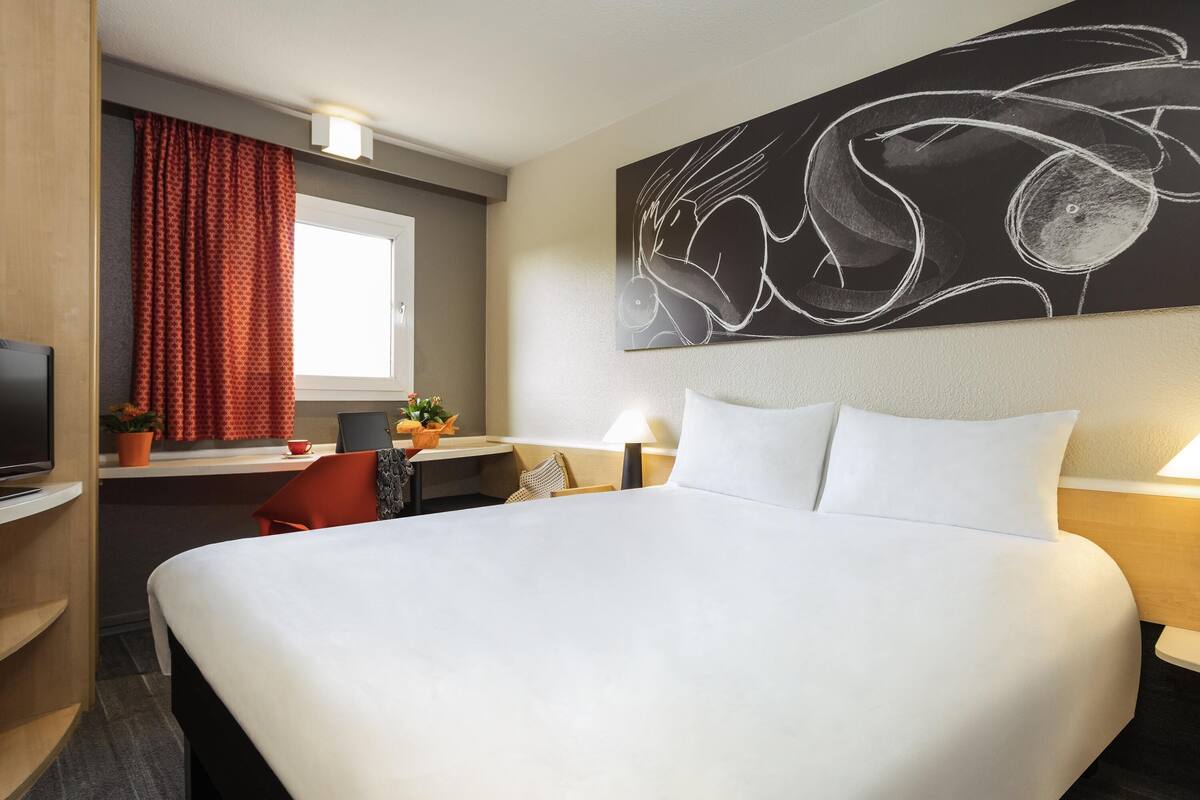 A hotel room with a large bed, a desk with a chair, a television, and a window with curtains.