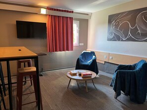 A room with a flat-screen TV mounted on the wall, a round table with a chair, and a sofa with a blanket. - ibis Paris Nord Villepinte parc des Expositions (Villepinte)