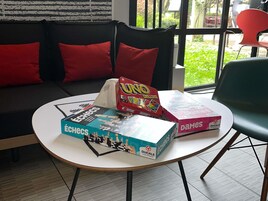Una habitación con una mesa redonda, dos juegos de mesa, un sofá con cojines rojos y una ventana que da a zonas verdes.